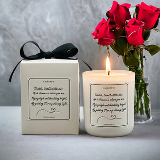 A beige candle with personalized text on the label, placed next to its white gift box adorned with a black ribbon and a vase of red roses in the background.
