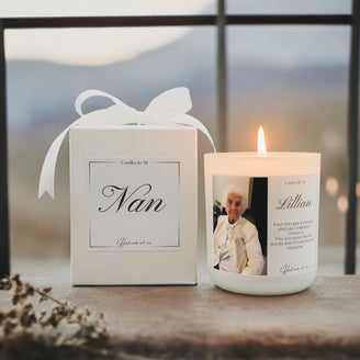 A white candle with a custom message label, placed next to its matte white gift box with a satin ribbon, on a wooden surface with a blurred landscape in the background.