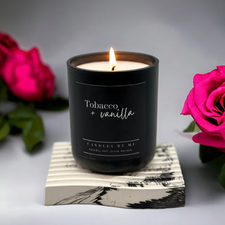 A black candle with a white label on it, branded 'Tabacco + Vanilla', placed on a coaster with a printed design.