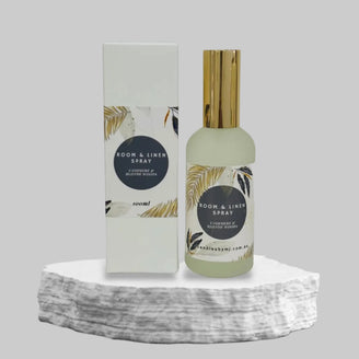 A room and linen spray bottle with a white label featuring a palm leaf design, and a gold-colored spray nozzle. The label includes text indicating the product name and scent notes.