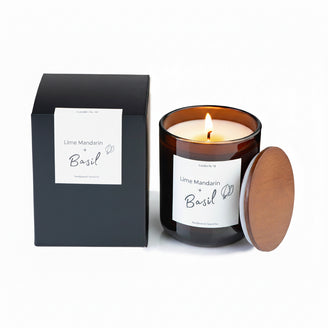 A black candle with a clear label reading 'Lime, Mandarin & Basil' alongside its packaging, which is also black with a simple design and a candle silhouette in the background.