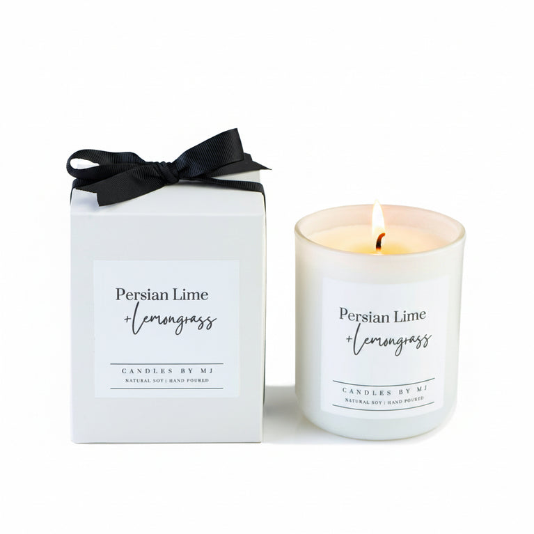 A white candle with a label reading 'Persian Lime Verbena' alongside its packaging, which is also white with a black bow on top.