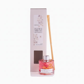 A 200ml bottle of SHEER LILY & WHITE ROSE reed diffuser with a silver cap and several dried flower reeds placed upright next to it.