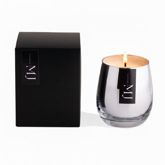A black candle with a clear glass base and a silver lid, alongside its black packaging that has the brand name 'MJ' on it.