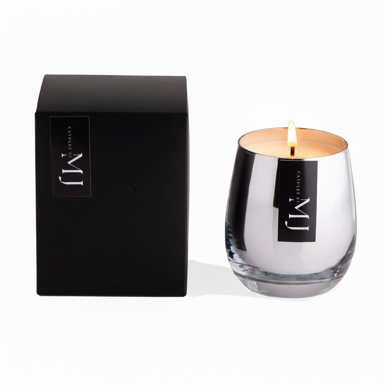 A black candle with a clear glass base and a silver lid, alongside its black packaging that has the brand name 'MJ' on it.