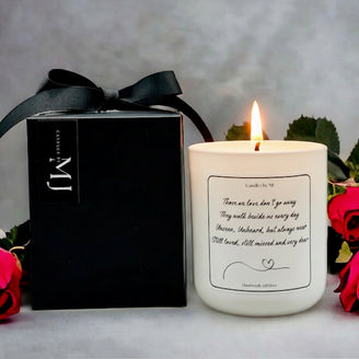 Candle with a message on it next to a black box with a bow, surrounded by flowers.