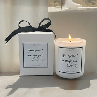 A custom message candle with a black label that reads 'YOUR SPECIAL MESSAGE GOES HERE' alongside a heart symbol, placed next to its white gift box with a black ribbon.